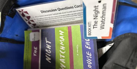 Blue and black tote bag. Inside the bag is the novel 'The Night Watchman', a set of Discussion Questions and a tag labeling the tote as: Book Kit-The Night Watchman